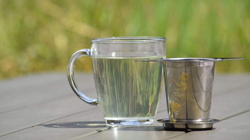 Image of a glass of honeysuckle tea with a stainless steel cup infuser holding the honeysuckle next to it, on a garden table with grass in the background.