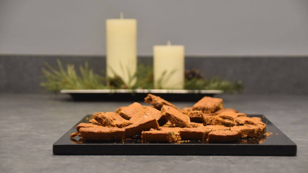 Image of a pile of spiced cookies on a black cutting board, with some Christmas decoration blurred in the background